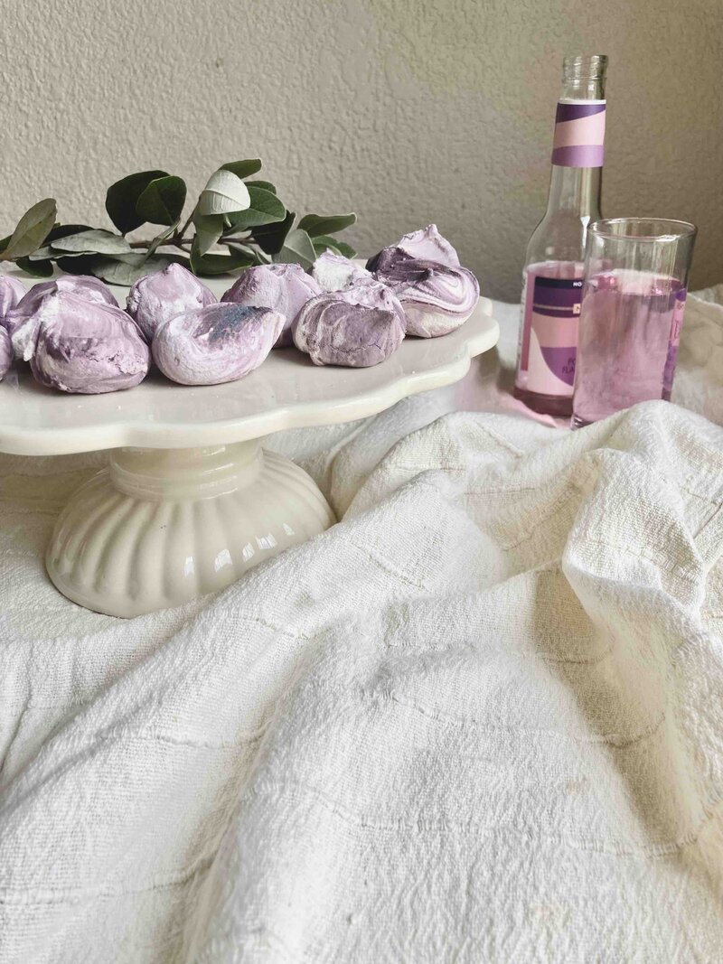 Purple hand piped meringues on a cake display, with a bottle of pink coloured juice.