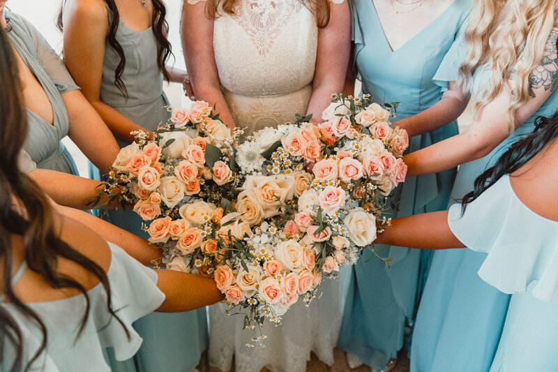 Bridesmaids in light blue dresses forming a heart shape with their hands around a beautiful bouquet of pastel roses at Ashton Gardens North in Houston, Texas, during Zach and Emily’s wedding.