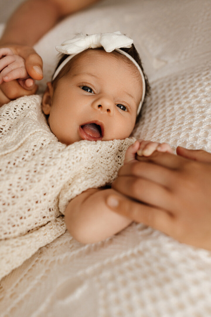Awake newborn baby girl with white bow lying on a textured blanket, gently holding family hands during a cozy in-home lifestyle session in Las Vegas by Angela Wolf Photography.