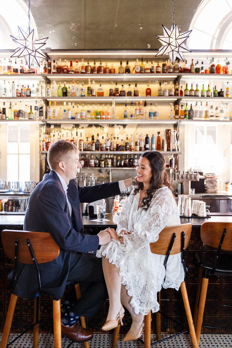 In an ambient Philadelphia bar, a couple laughs across a row of cocktail glasses and exposed brick. The bride-to-be wears a romantic lace dress with sleeves while her fiancé rests his arm on the back of her chair. Warm light from industrial fixtures and a fully stocked backlit bar bring a cozy, celebratory energy to these Philly engagement photos.