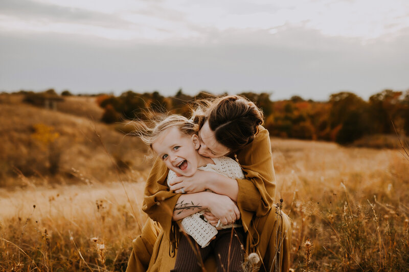 A mom holding her daughter from behind giving her a hug and laughing in a tall grassy field in Fort Worth, Texas