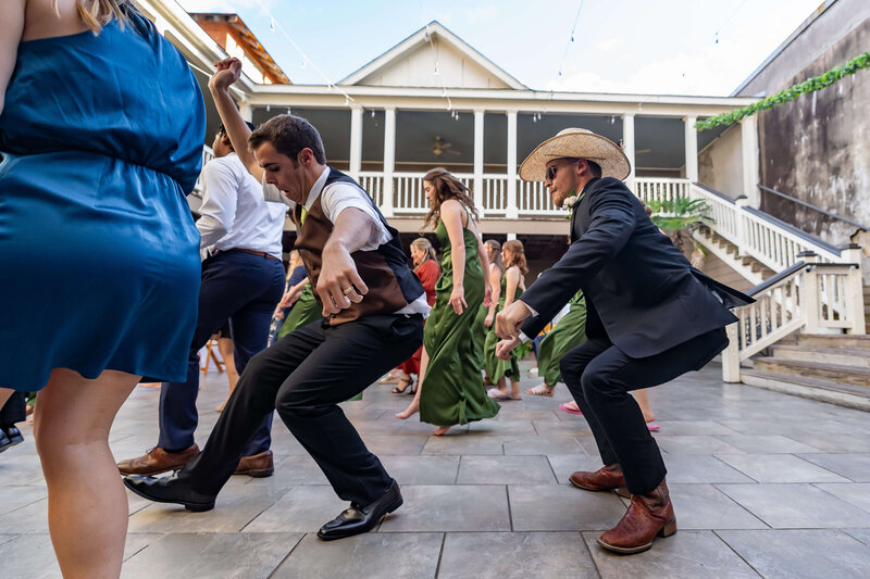 A group of people dressed in formal attire dancing enthusiastically outdoors on a tiled patio. One man wears a cowboy hat and boots while others join the fun in suits, dresses, and green gowns. A staircase is in the background.