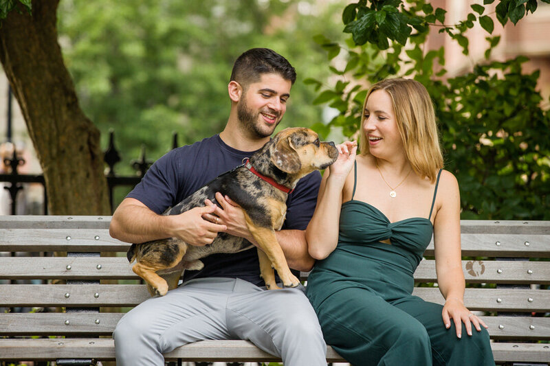 An engaged couple giving their dog a treat during an engagement session in Boston, capturing a sweet and joyful moment that highlights their bond with their furry companion amid the city's charm.