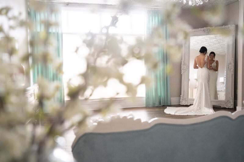 Bride in elegant white lace wedding dress with train being helped by bridesmaid in front of ornate white mirror in bright, airy bridal suite at the White Room venue in historic downtown St. Augustine