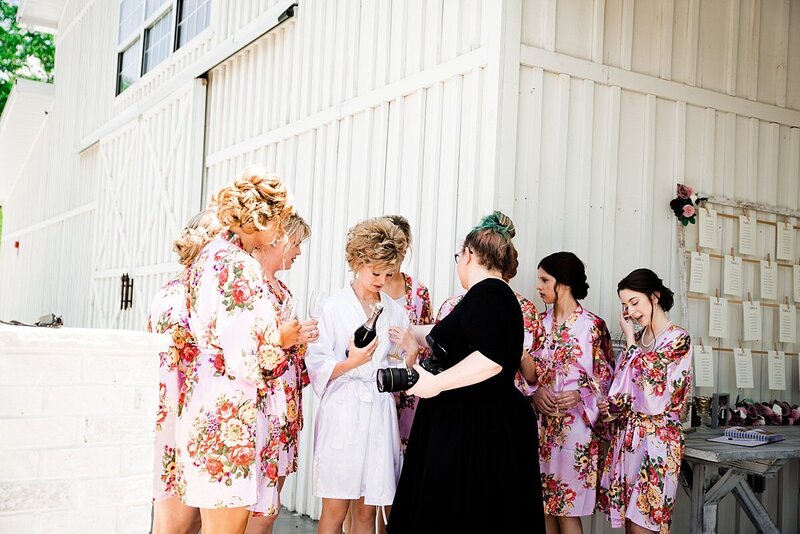 Bridesmaids wearing floral robes standing outside getting ready to pop champagne, Mahlia is showing them how to set up for the best photo