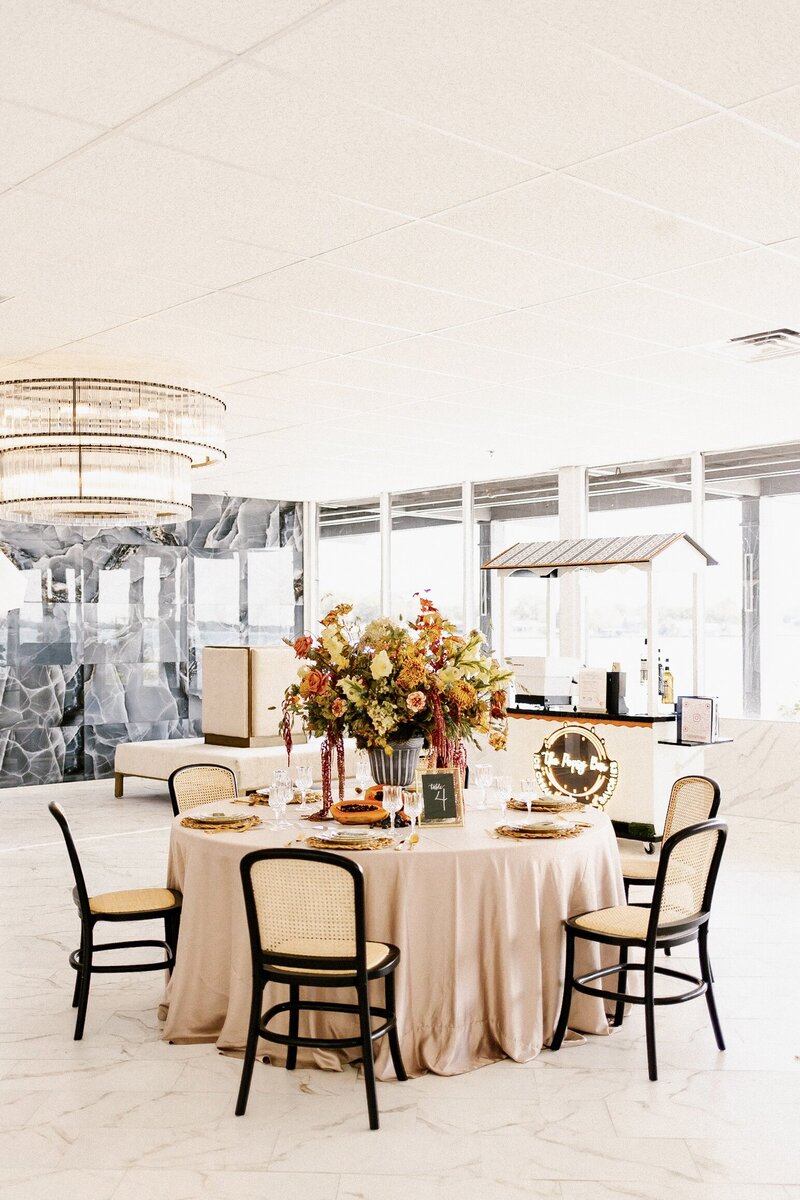 Blush-linen round table with black cane-back chairs, abundant fall-toned centerpiece, and bar backdrop in a bright modern venue.
