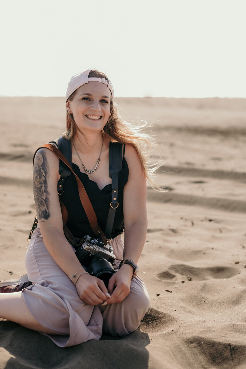 Karin Busch, photographer, is sitting on the beach in the sand, smiling.