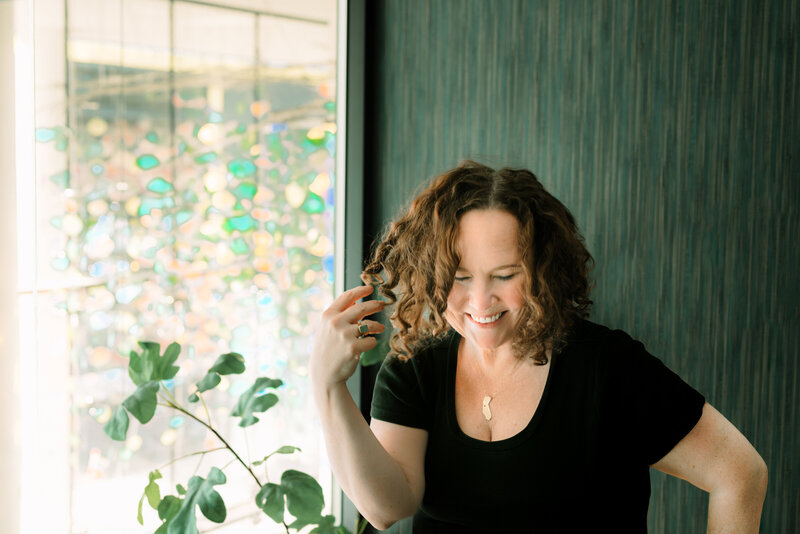 A portrait of the photographer  laughing and touching her shoulder length curly brown hair.  She  is wearing a black scoop neck t-shirt with a gold necklace in the shape of California.  standing in front of a wall with teal fabric wallpaper and beside a plant that is in front of a window with colorful reflections in the background