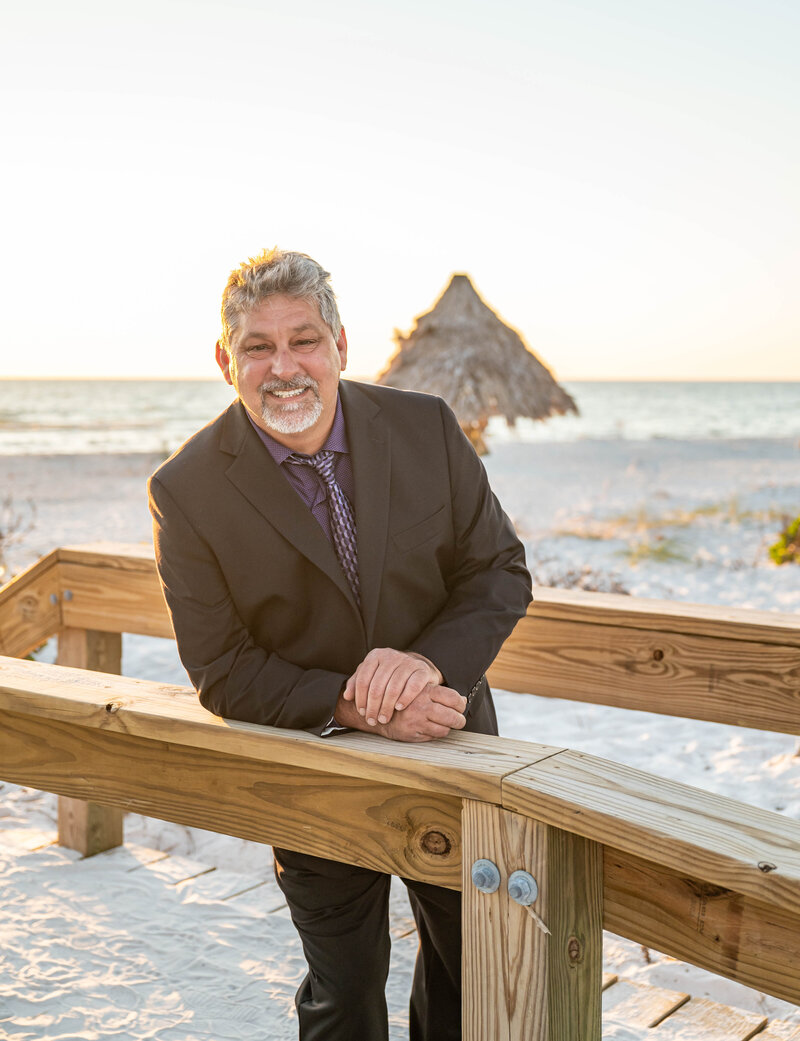 Photo of John Corbenllini smiling in a suit leaning against a beach walkway with the ocean in the background