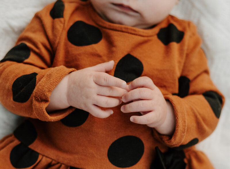 Milestone photo of baby girls hands while laying down wearing an orange and black polka dotted dress.