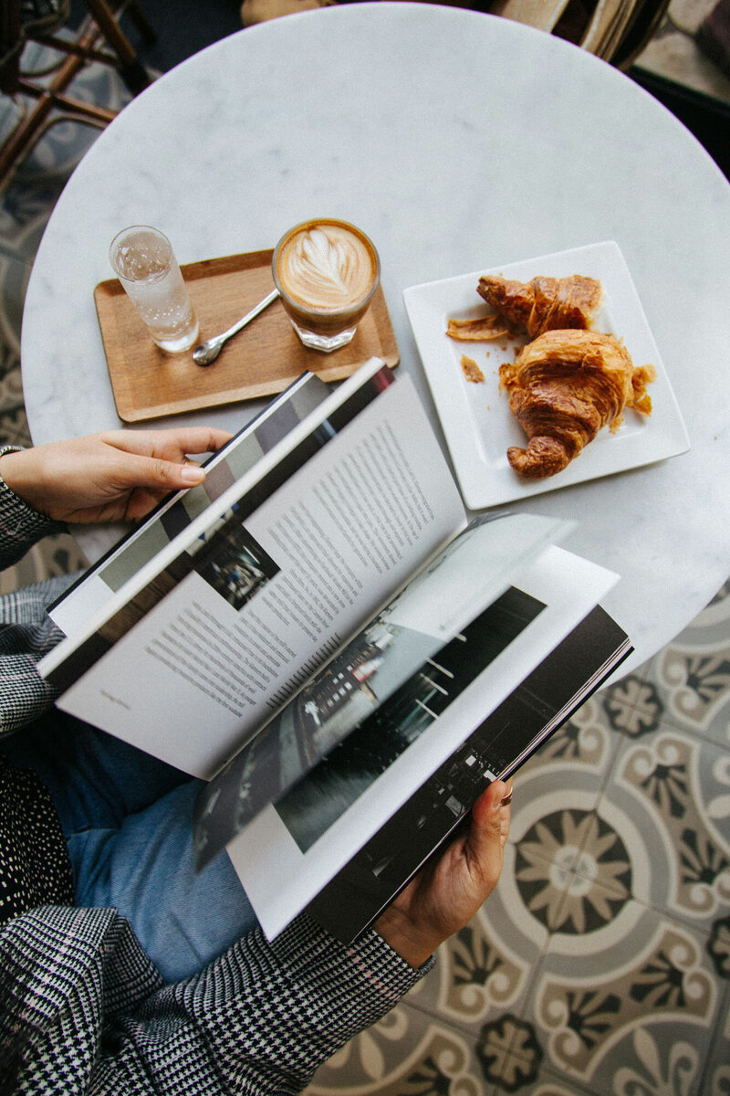 Person reading a book next to a plate of food and a cup of coffee