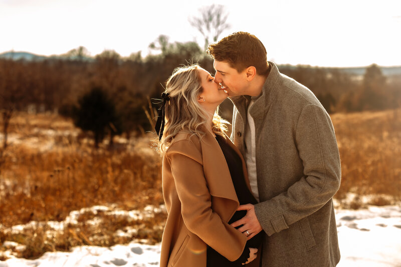 A man and women are standing in a field with tall grass with trees in the background, and there's snow on the ground. They're just about to kiss in a loving embrace while the man holds the women's pregnant belly.