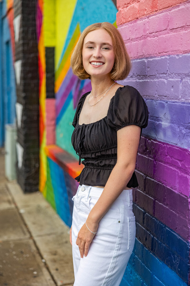 A female high school senior poses next to a painted mural brick wall in Ann Arbor, MI.  She is wearing a black shirt and white pants