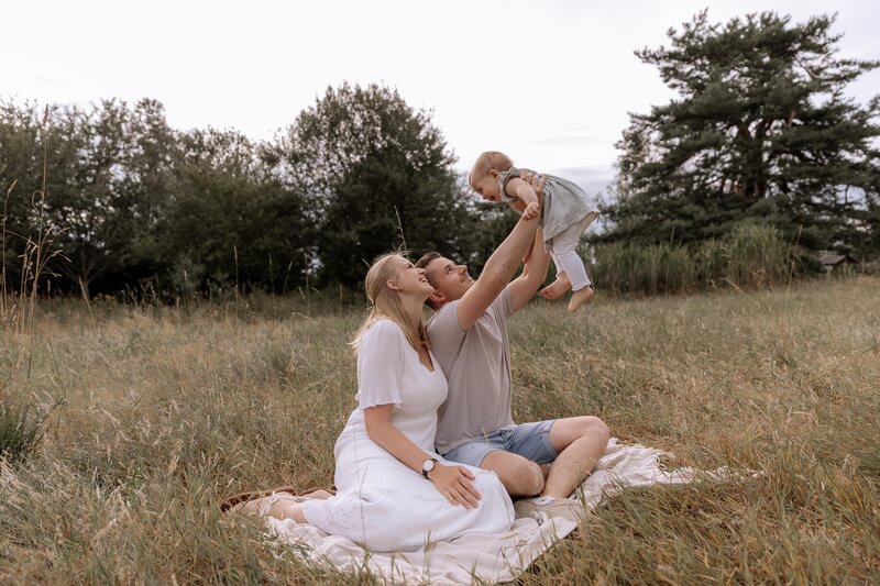 Mutter und Vater sitzen auf einer Decke und halten Kleinkind in die Luft