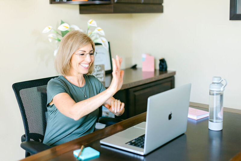 Lora Ulrich waving at the camera on her laptop while sitting at her office