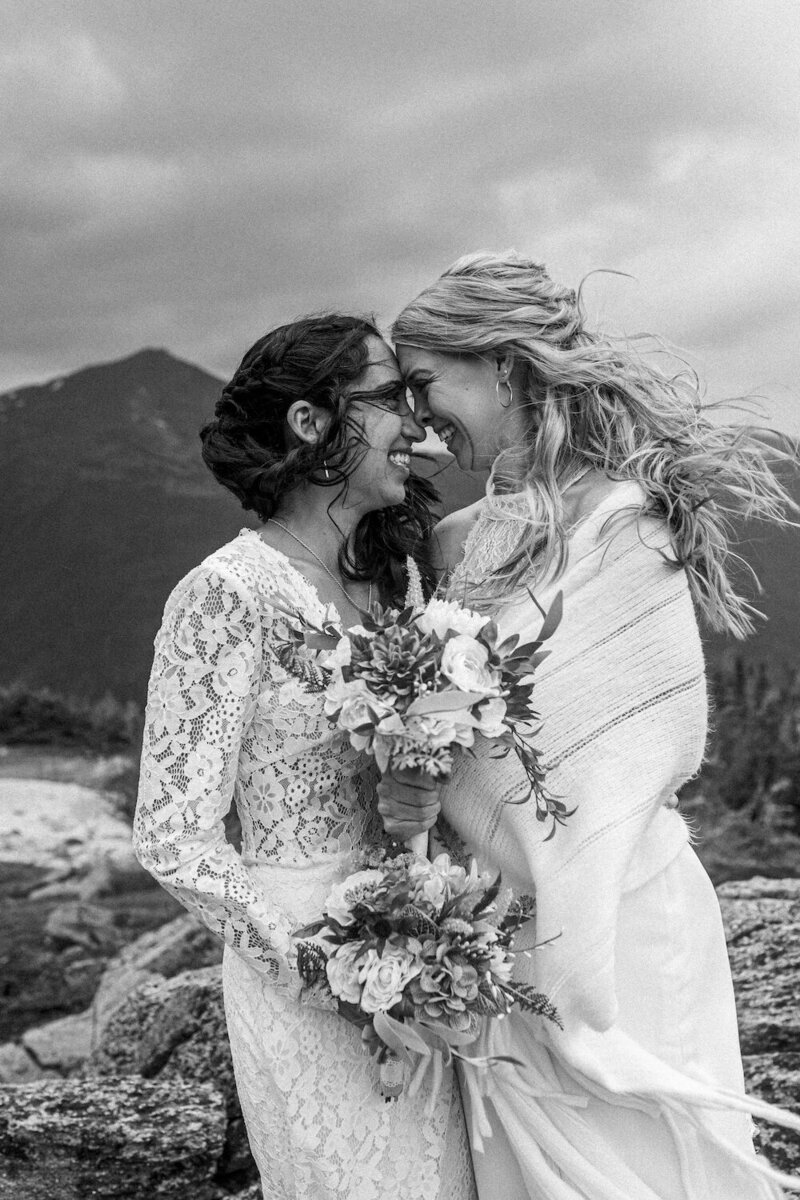 Two brides in wedding dresses, holding flowers and touching foreheads as the wind blows their hair during their elopement on Mount Washington.