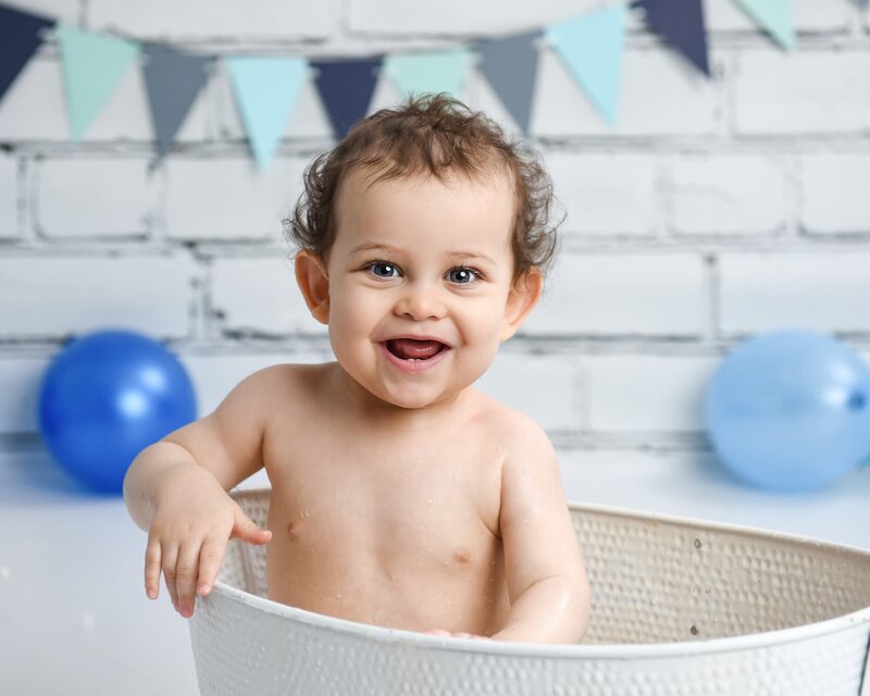 A smiling baby boy sitting in a white tub, with blue balloons and pastel-colored bunting on a white brick wall backdrop.