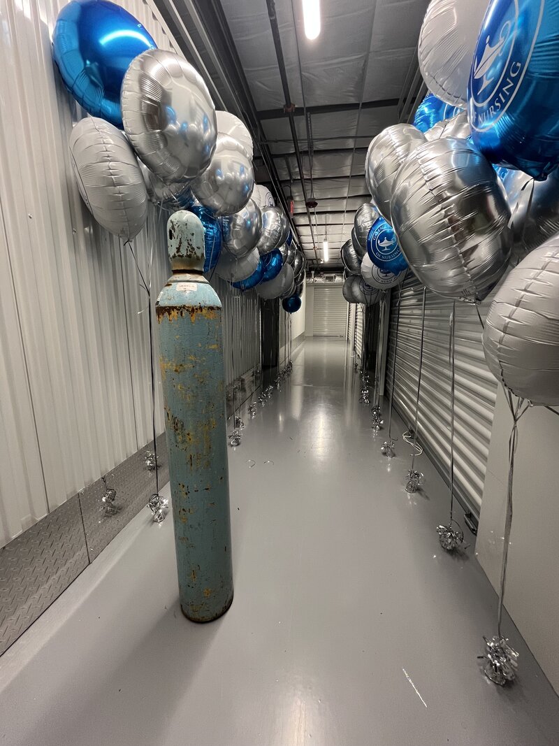 Helium tank and silver, blue, and white helium balloons with Galen College of Nursing on them positioned behind the helium tank in a long hallway. 