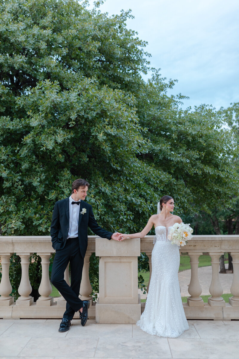 A bride and groom holding hands on a balcony, the bride in a sparkling white dress and the groom in a sleek tuxedo, sharing a quiet moment.