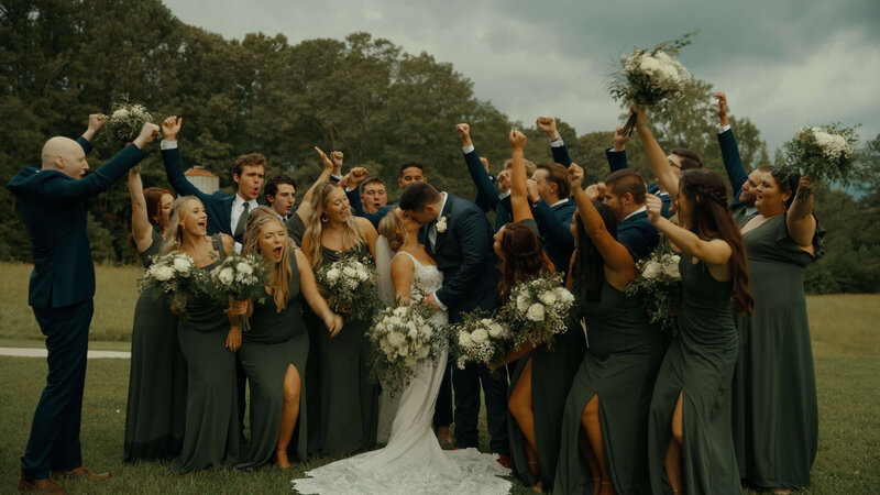 The groom leaned in to kiss the bride on the green lawn, surrounded by bridesmaids and groomsmen.