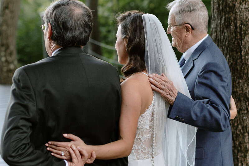 A bride in a white gown and veil stands between two men, showcasing a heartfelt moment of support and connection during a ceremony.