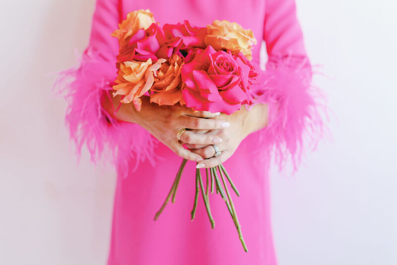 Woman in bright pink dress with feather sleeve cuffs holding a pink and orange bouquet in front of her
