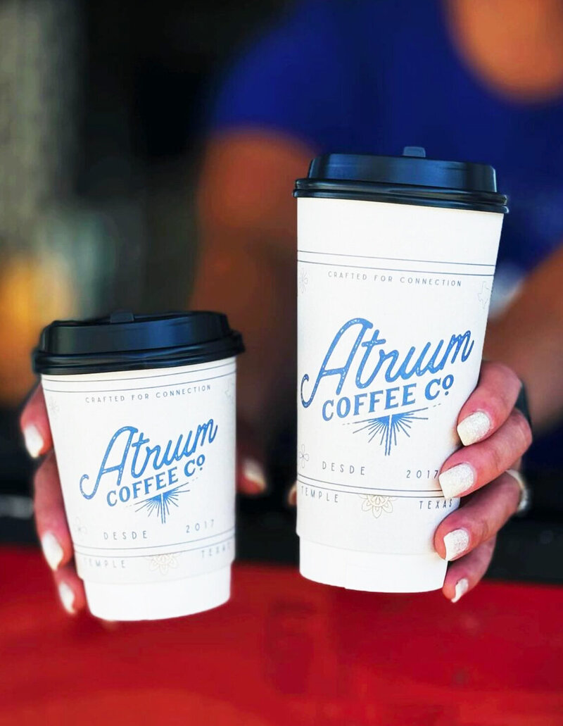 Close-up of two hands holding takeaway coffee cups with black lids, labeled "Atrium Coffee Co." The person has white painted nails, and a blurred background creates a focus on the cups, capturing the essence of Temple, Texas's favorite coffee shop.