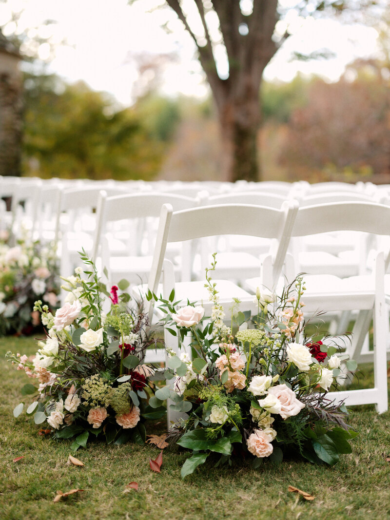 November wedding at Graylyn Estate, designed and planned by Charming Carolina — ceremony aisle lined with blush, burgundy, and ivory floral ground arrangements with soft greenery in the White Garden.