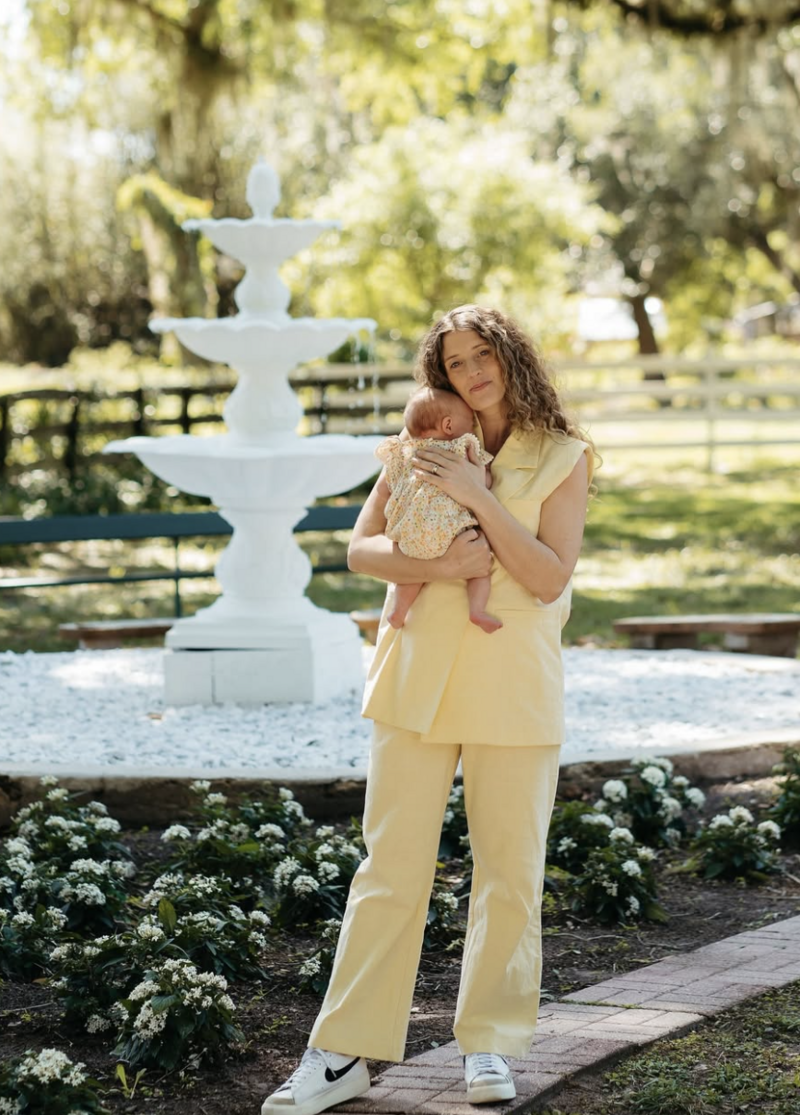 Mother in yellow holding her baby near the white fountain at Uliana Estate, a serene and family-friendly wedding venue in Lakeland Florida.
