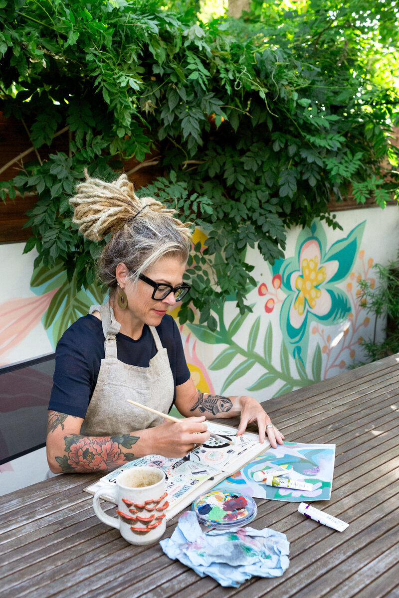 Calgary brand storytelling photoshoot of a woman with dreadlocks, black glasses, and tattooed arms painting in an outdoor creative space. She wears a linen apron over a navy shirt and is focused on her sketchbook, surrounded by paint supplies and a textured ceramic mug. The backdrop features lush greenery and a vibrant, hand-painted floral mural, creating an inspiring and artistic atmosphere.