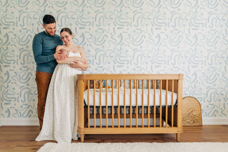 Mom and Dad standing next to crib with blue swirled wallpaper behind them holding newborn baby