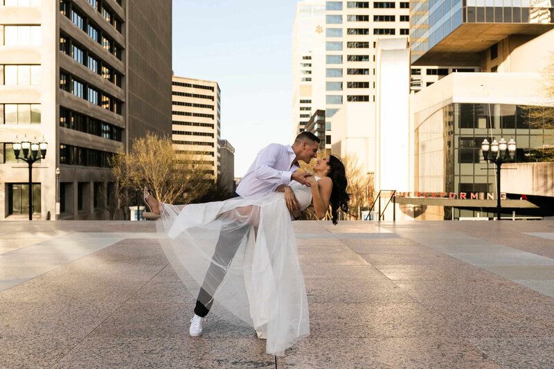 In the middle of a city plaza, a couple shares a dramatic dip as golden Northeastern light hits the glass and concrete buildings around them. The bride wears a white tulle skirt that floats mid-air as she’s lifted by her partner, who wears white sneakers and a crisp button-down. T