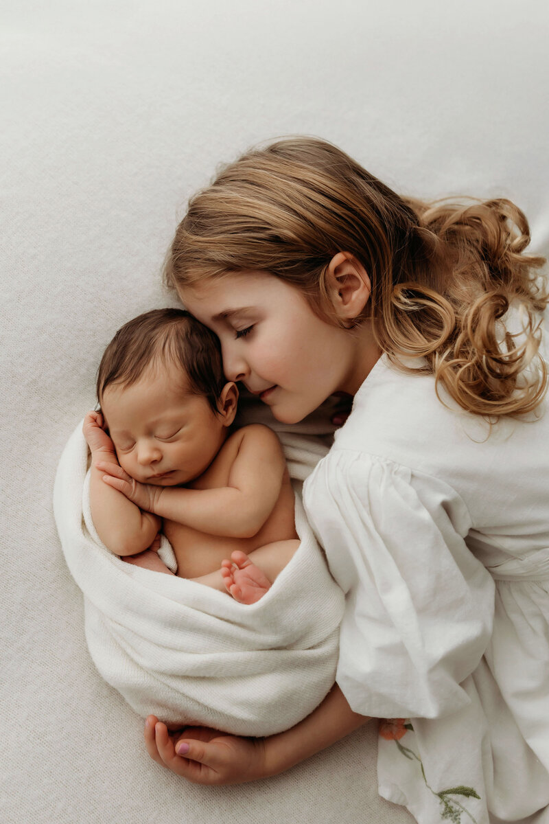  A newborn baby sleeps wrapped in blankets next to her big sister who is snuggling close on a bed of cozy blankets for her newborn photo session with Vancouver Washington Photographer