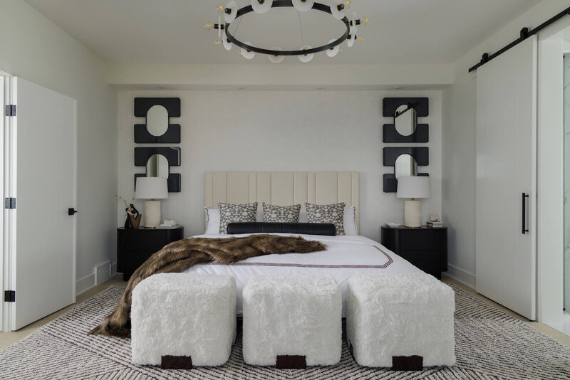 A primary bedroom with fur ottomans and soft linens paired with black accent mirrors and lighting. 