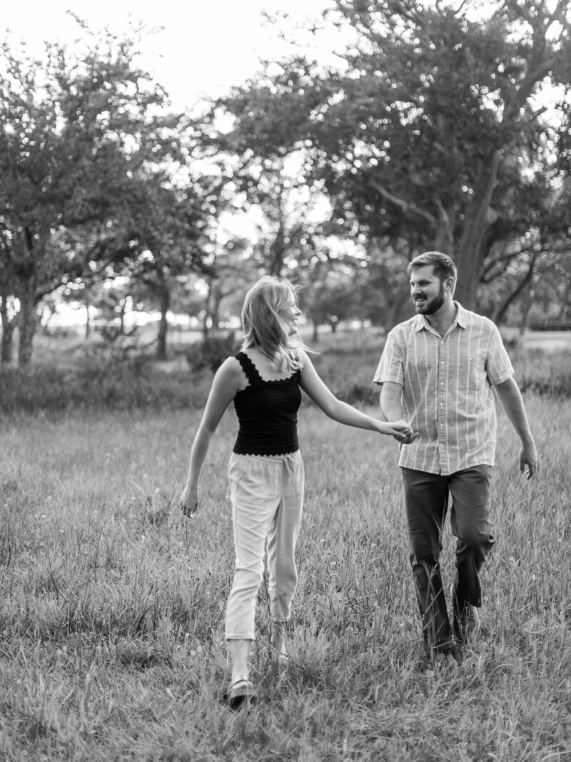 Outdoor engagement session. Couple holding hands as they are walking through the grass and looking at eachother.