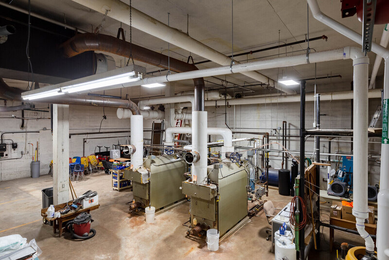 Engineering boiler room with green industrial boilers, overhead white insulated piping, and fluorescent lighting in a large mechanical space.