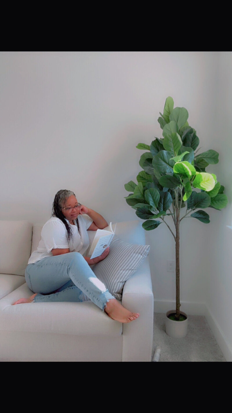 Letitia Hopkins sitting on a cream couch reading a book, embodying personal growth, self-love, and Main Character Energy as a confidence coach helping women transform their lives.