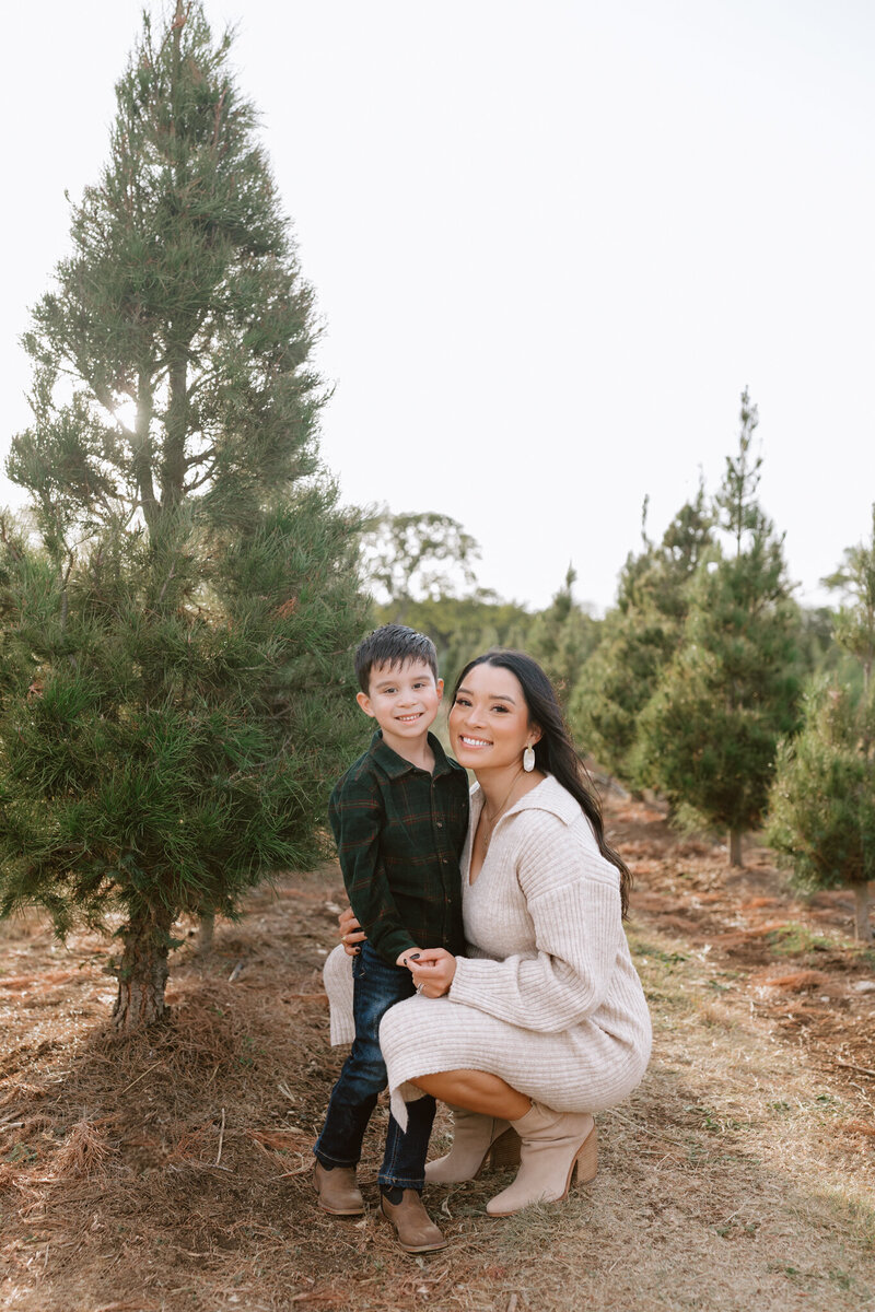 A mom in a cream flowy dress holds her toddler son wearing a cream button up. They are walking through a grassy San Antonio field.