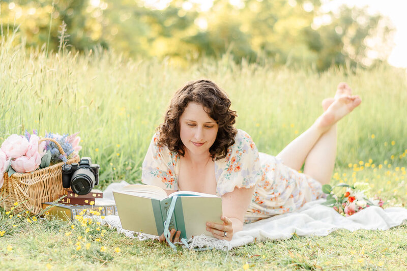 A woman, wearing a floral dress, lays on a white blanket reading a book. Around her are other flowers, a  basket, and a stack of books topped with a digital camera.