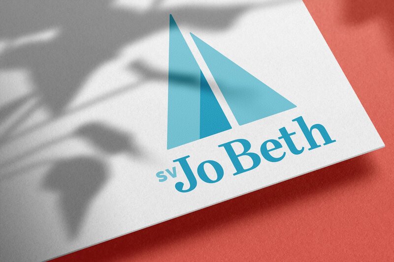 A blue logo featuring triangular sails above the name "SV Jo Beth" printed on a white piece of paper sitting on a coral background