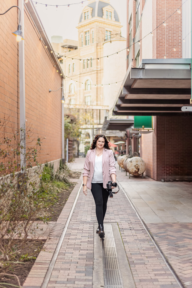Jennifer in pink jacket holding camera and walking during branding session at The Pearl in San Antonio, Texas.