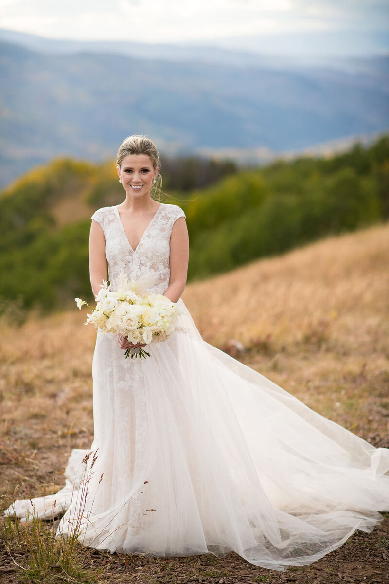 Photo of bride in a flowing toile dress posing and smiling, holding a white bouquet on a hilltop.