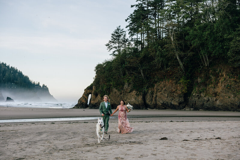 A bride wearing a gorgeous floral gown and a groom in a green suit run holding hands while their dog runs ahead on the Oregon Coast PNW Elopement. | Erica Swantek Photography