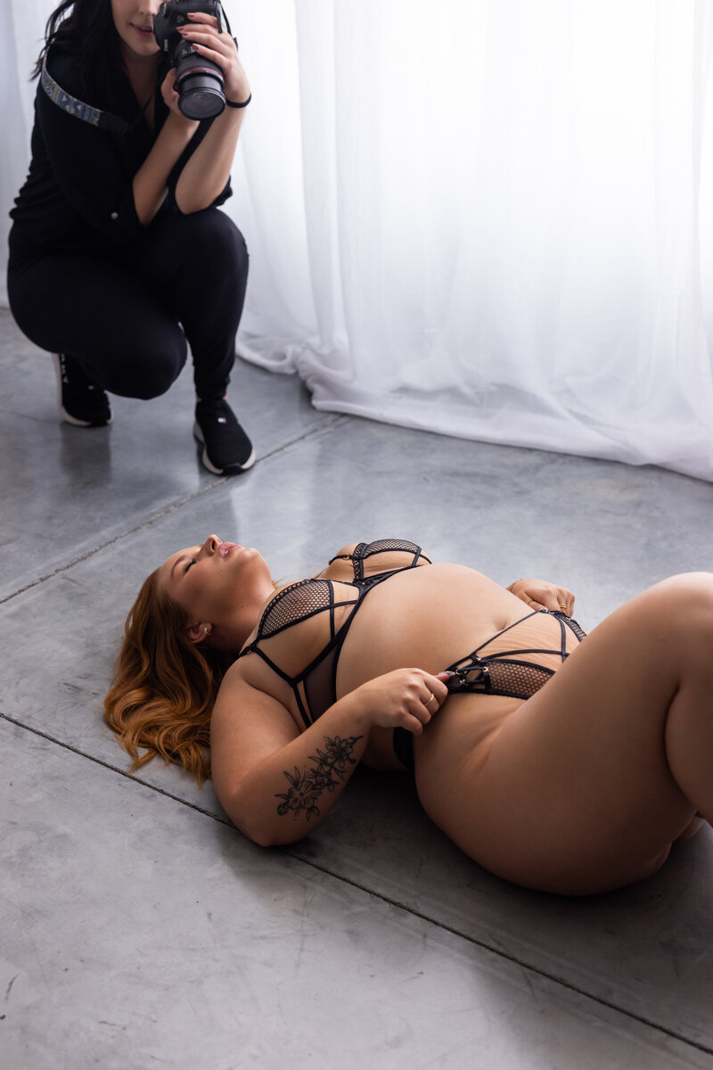 A woman in her 20s is kneeling on a bed with a black lace bodysuit while biting her thumb