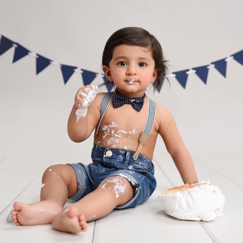 A baby boy wearing denim overalls, sitting on a white chair and reaching out playfully, with a colorful bunting and "ONE" letters in the background