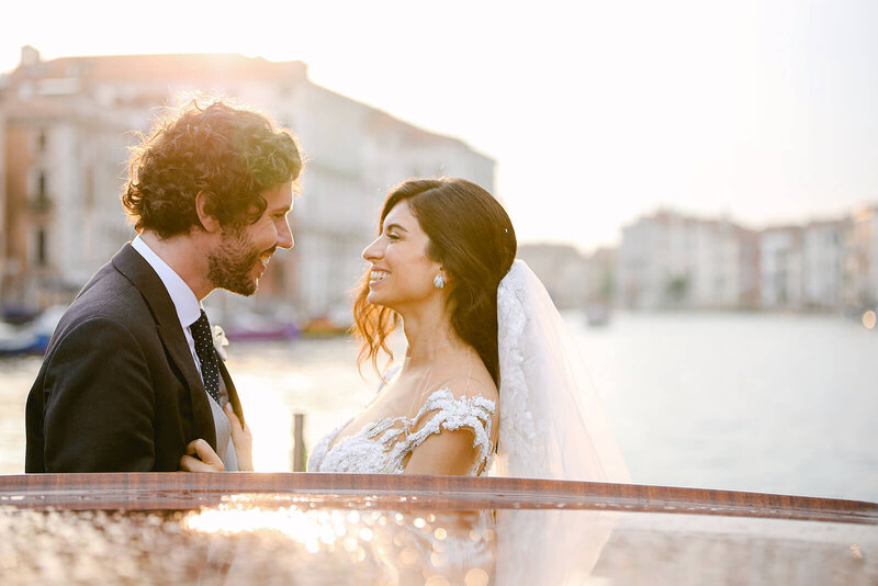 Couple just married looking each other, smiling happily. They are on a taxi boat in Venice at sunset.