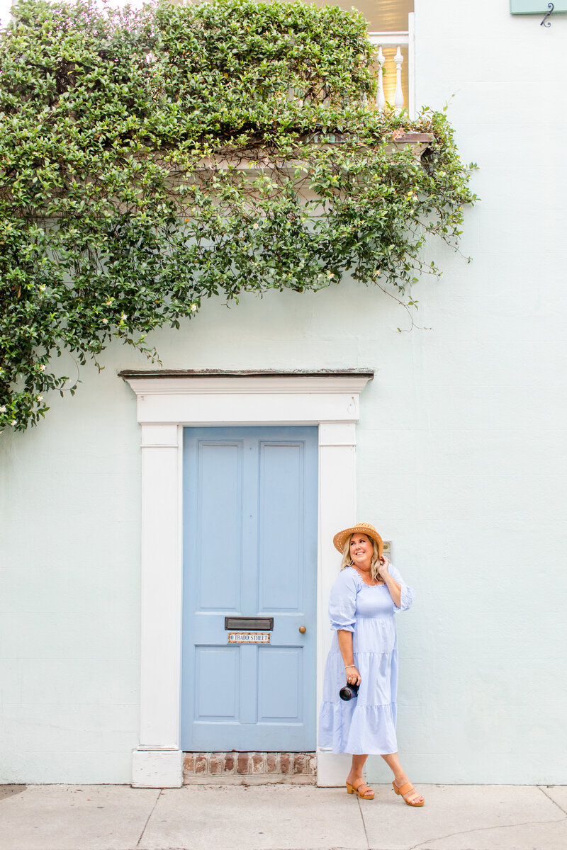 photographer posing in front of blue door in Charleston, South Carolina with cute hat and holding camera