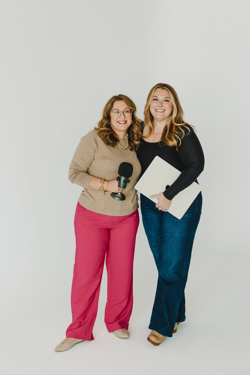 melissa and corry standing with a white background holding a microphone and laptop smiling at camera