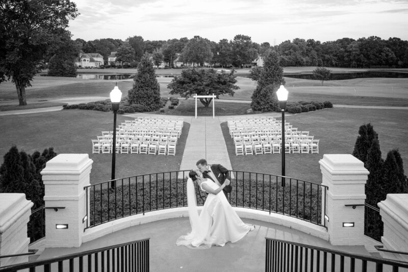 Black and white wedding photo of a bride and groom sharing a romantic dip kiss on the terrace at Providence Country Club in Charlotte, NC, with the outdoor ceremony space and golf course in the background.