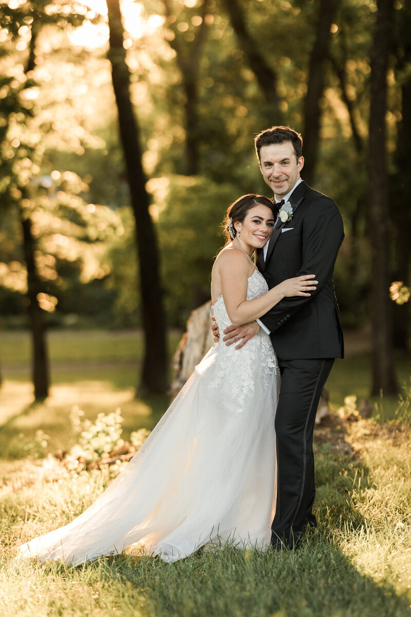 white groom in a black tux holding a white brunette bride with her head on his chest in a forest at sunset both looking at the camera smiling